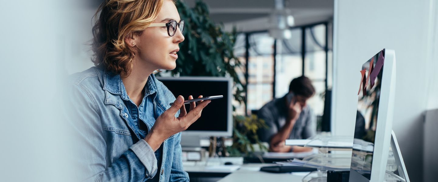 Junge Frau sitzt am Schreibtisch, schaut auf Ihren Bildschirm und hält ihr Smartphone in der Hand.